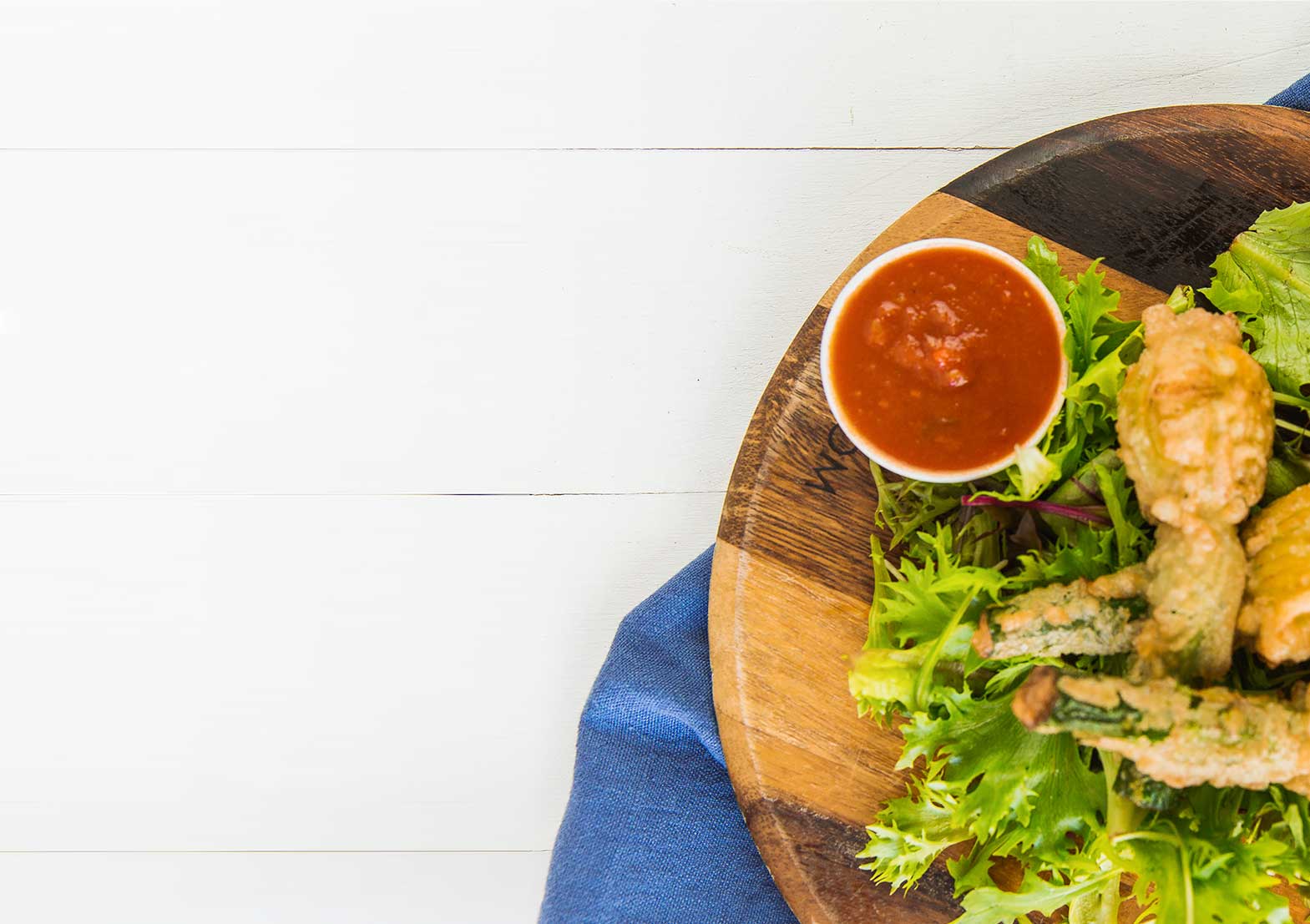 Fried Zucchini Blossoms with Tomato Dipping Sauce Partial view of a wooden serving board with fresh greens, fried zucchini blossoms, and a small bowl of red tomato dipping sauce.